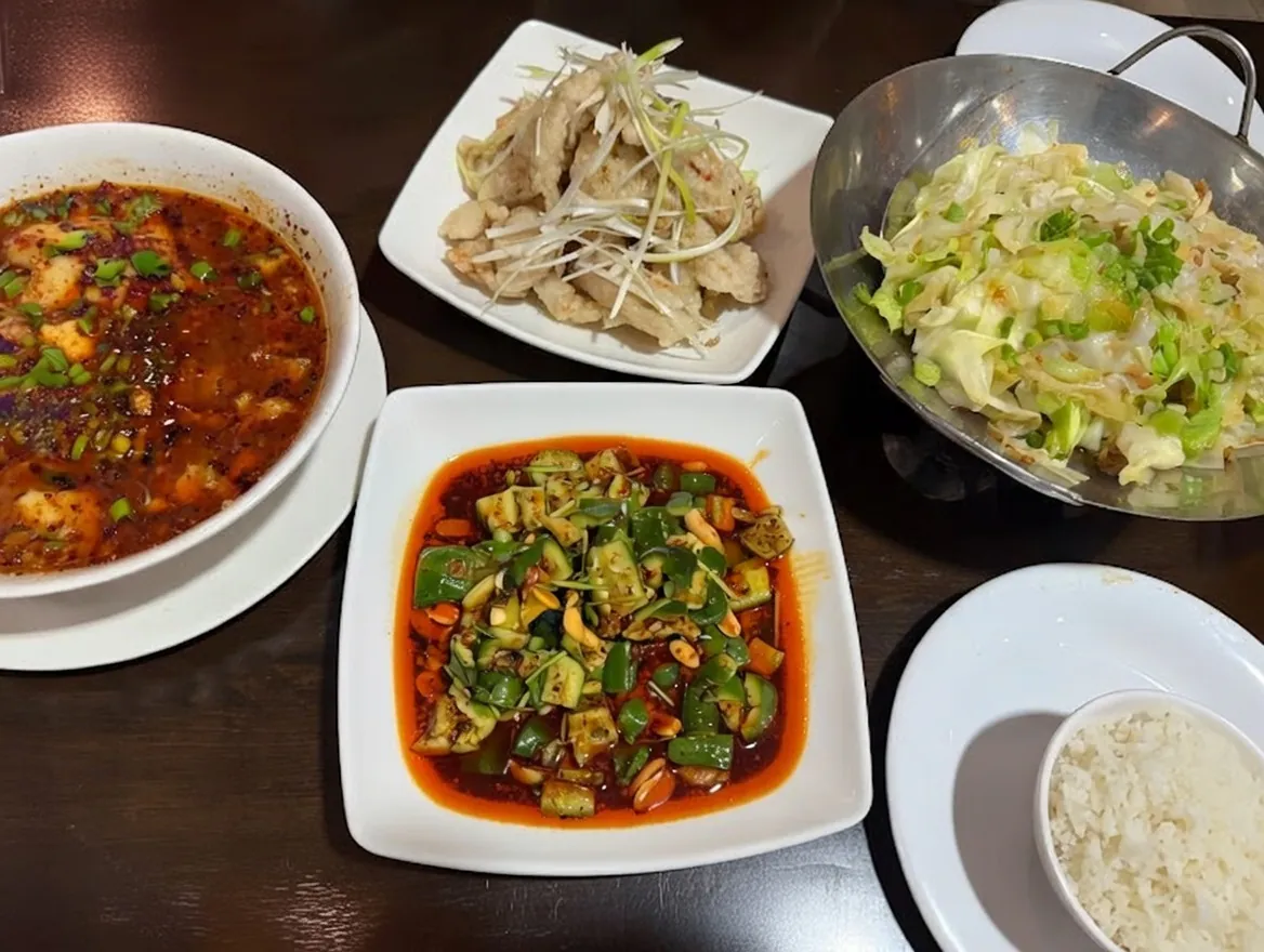 A table spread featuring four Chinese dishes: a spicy tofu soup, stir-fried cabbage, a cold cucumber salad, and a plate of chicken, served with rice.